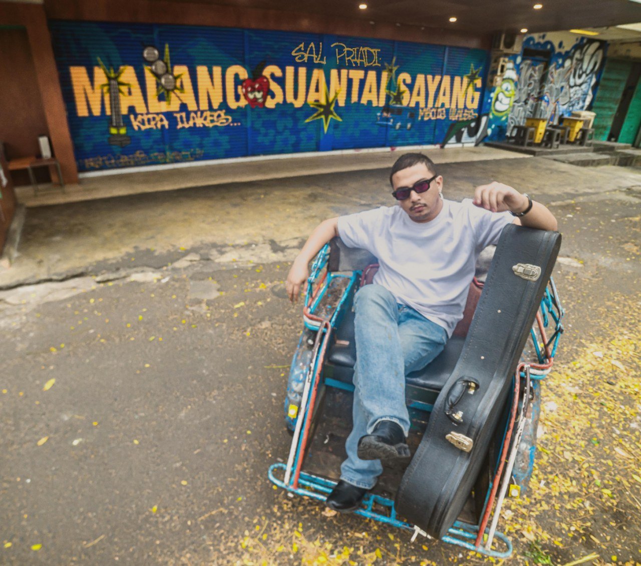 Sal Priadi berpose di becak dengan latar belakang mural judul lagu terbarunya